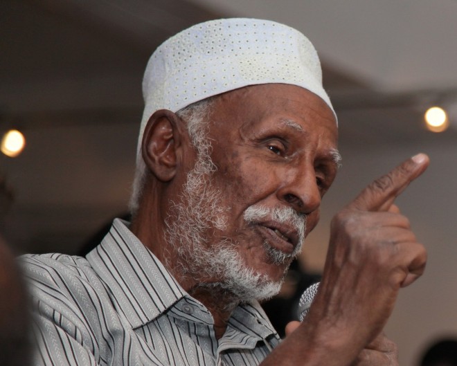 Elderly man in white Islamic skull cap (kufi) and grey and white striped collared shirt pointing up with right index finger.
